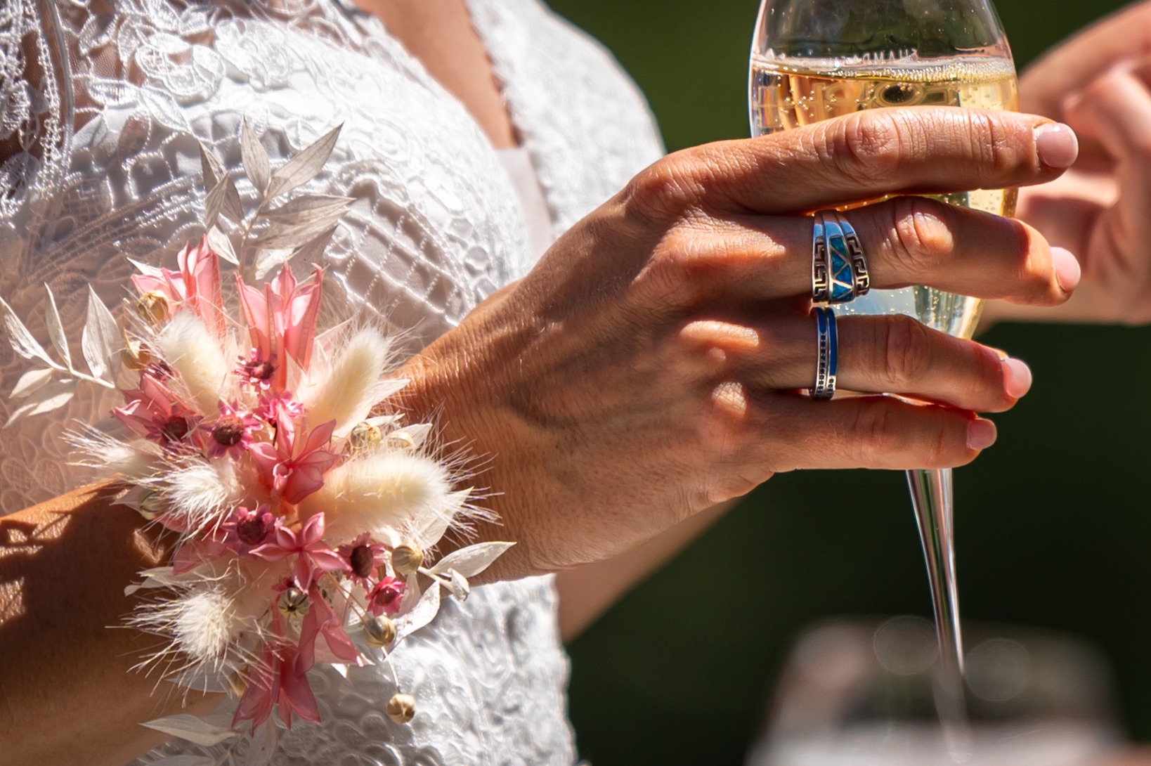 Détail d'une main de mariée tenant un verre de champagne avec une boutonnière florale.