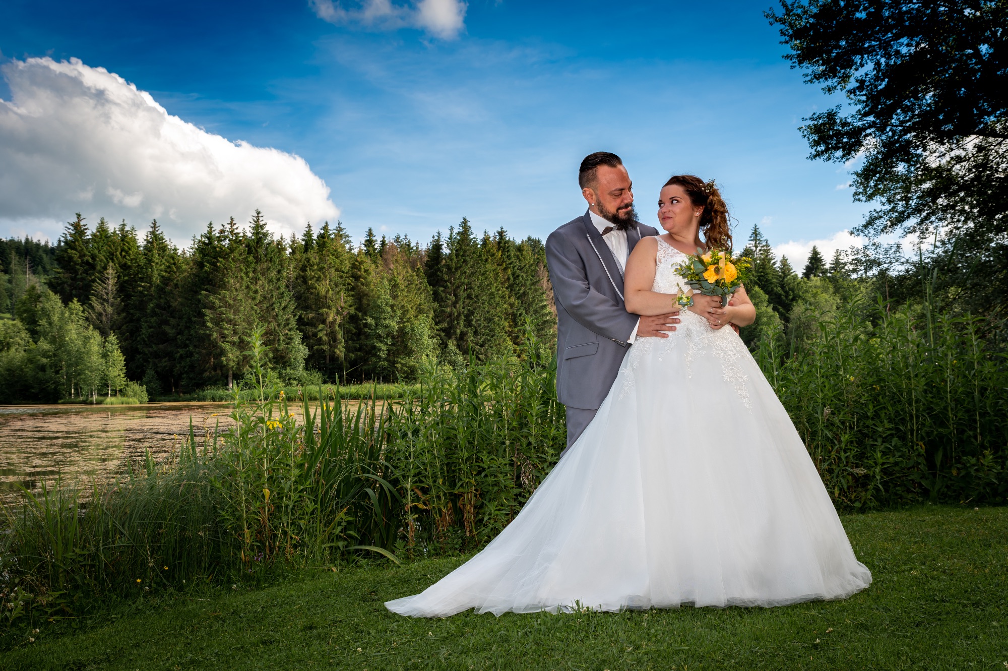 Couple de mariés posant au bord d'un lac, entouré de verdure.