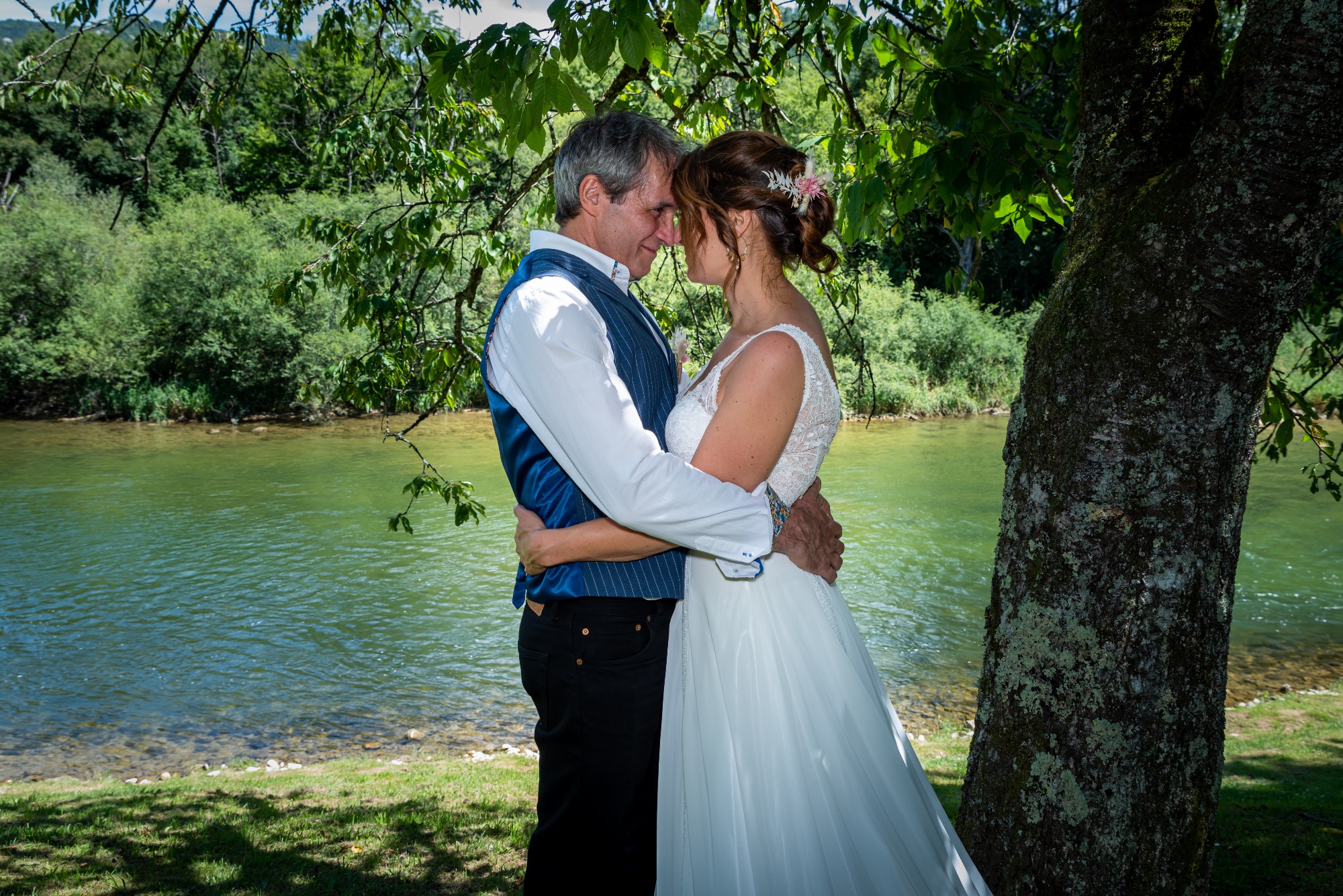 Couple de mariés s'embrassant au bord de l'eau, sous un arbre.